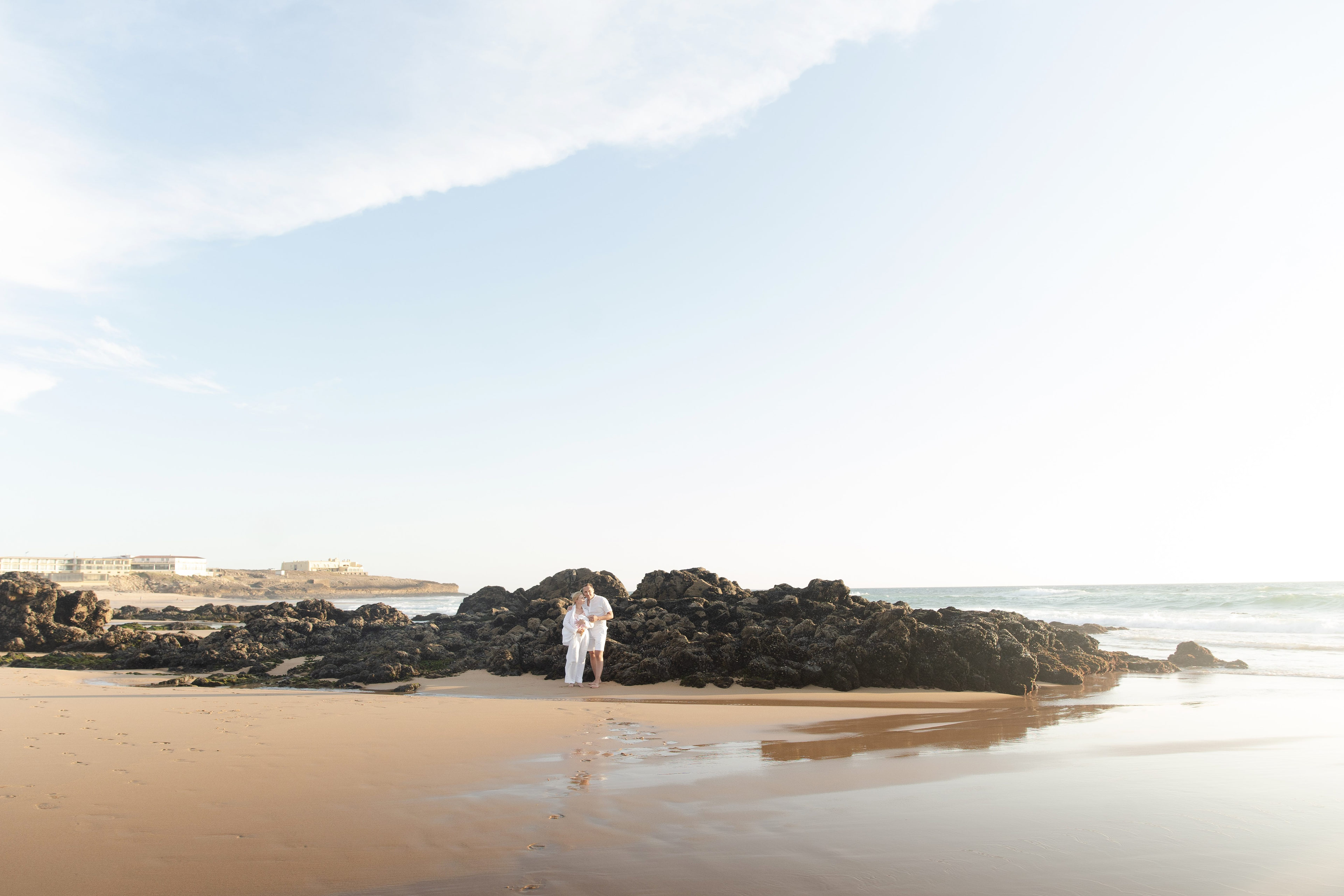 Family dressed in white standing in front of dark rocks on the wet sand at Guincho beach, Cascais Family dressed in white standing in front of dark rocks on the wet sand at Guincho beach, Cascais