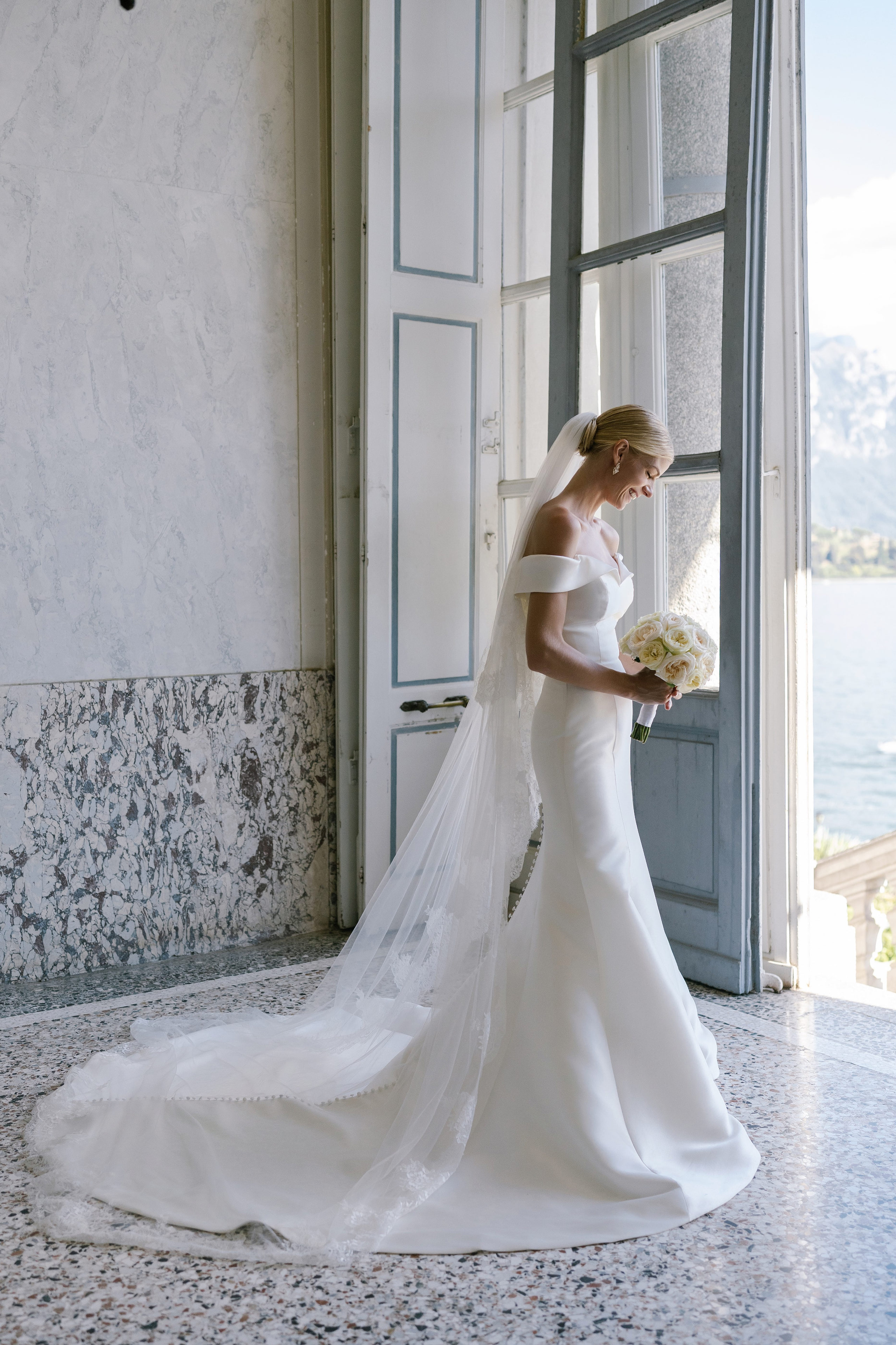 Bride in a fitted off-the-shoulder gown holding a white rose bouquet by a window at Villa Carlotta, Lake Como. Bride in a fitted off-the-shoulder gown holding a white rose bouquet by a window at Villa Carlotta, Lake Como.