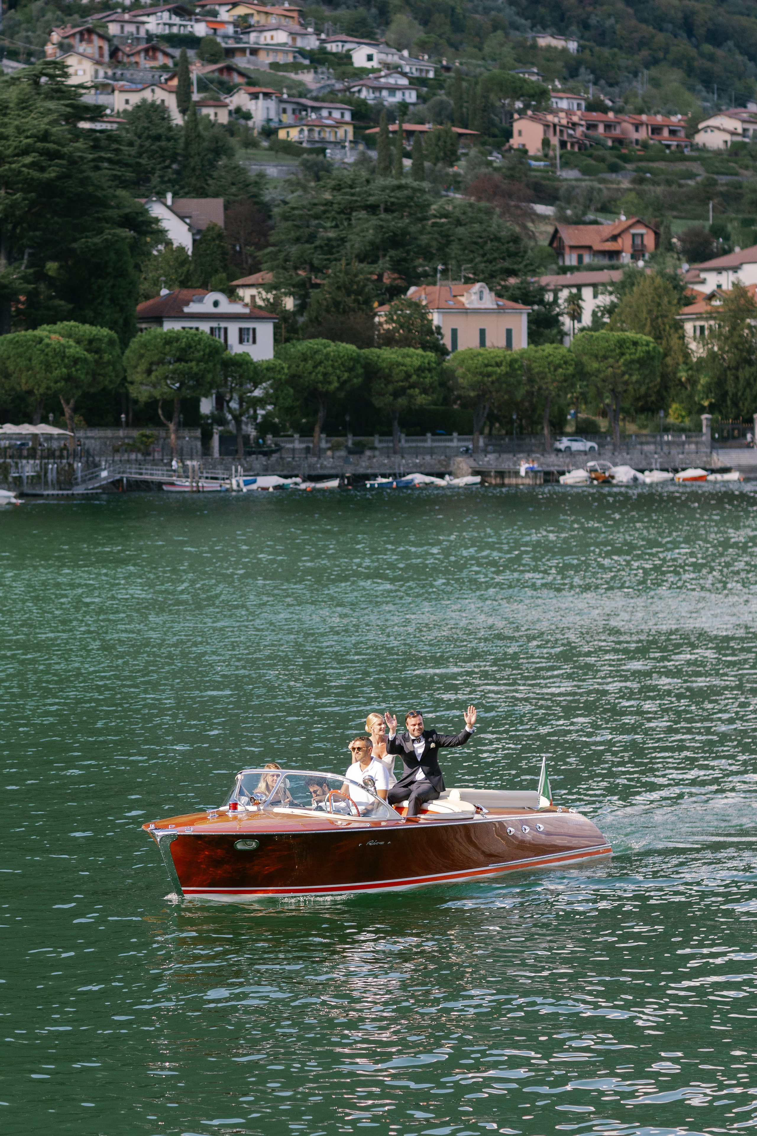 Bride and groom waving from a classic wooden speedboat on Lake Como after their Villa Carlotta wedding. Bride and groom waving from a classic wooden speedboat on Lake Como after their Villa Carlotta wedding.