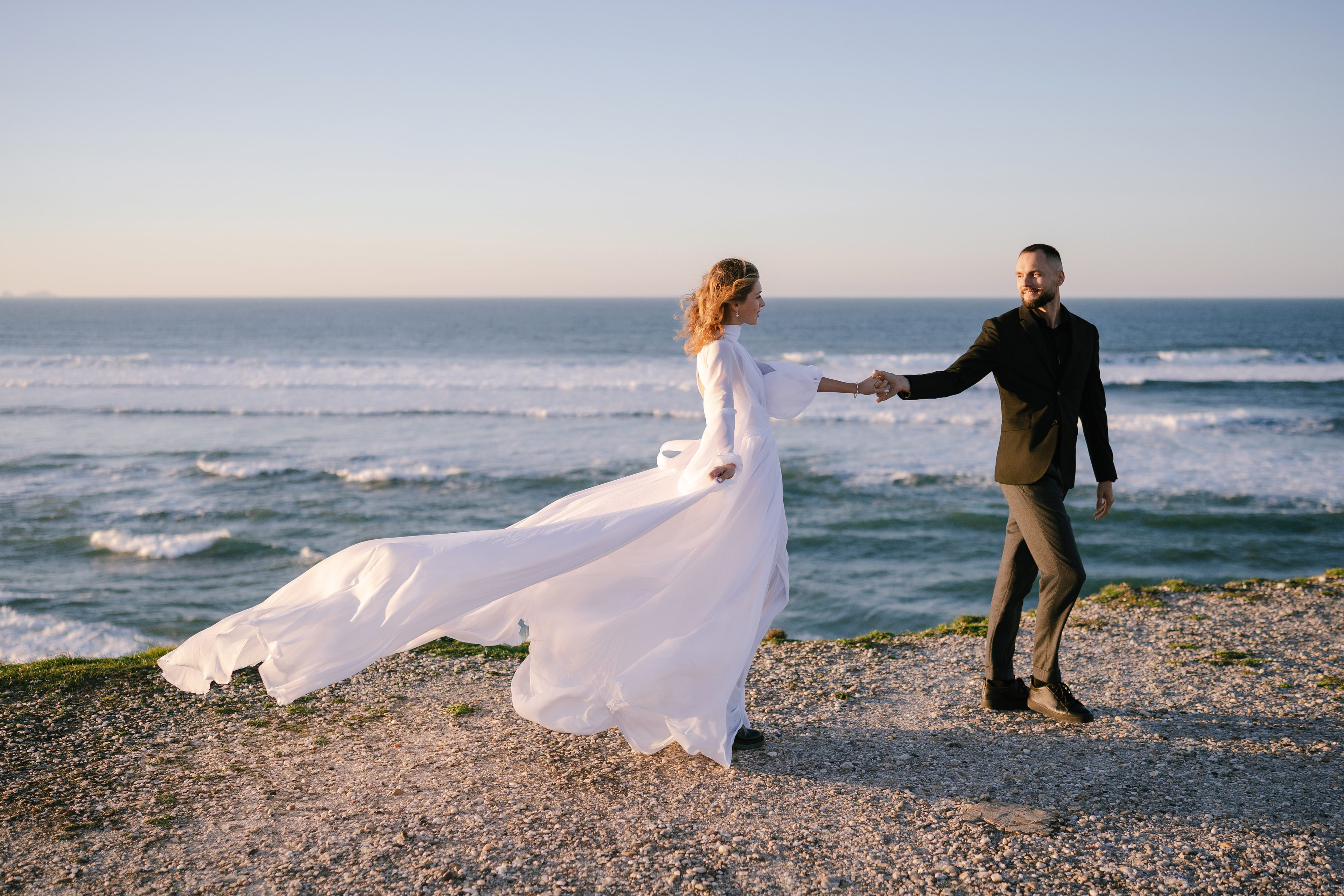 Bride and groom holding hands on the shore at Praia del Rey, her gown flowing in the wind